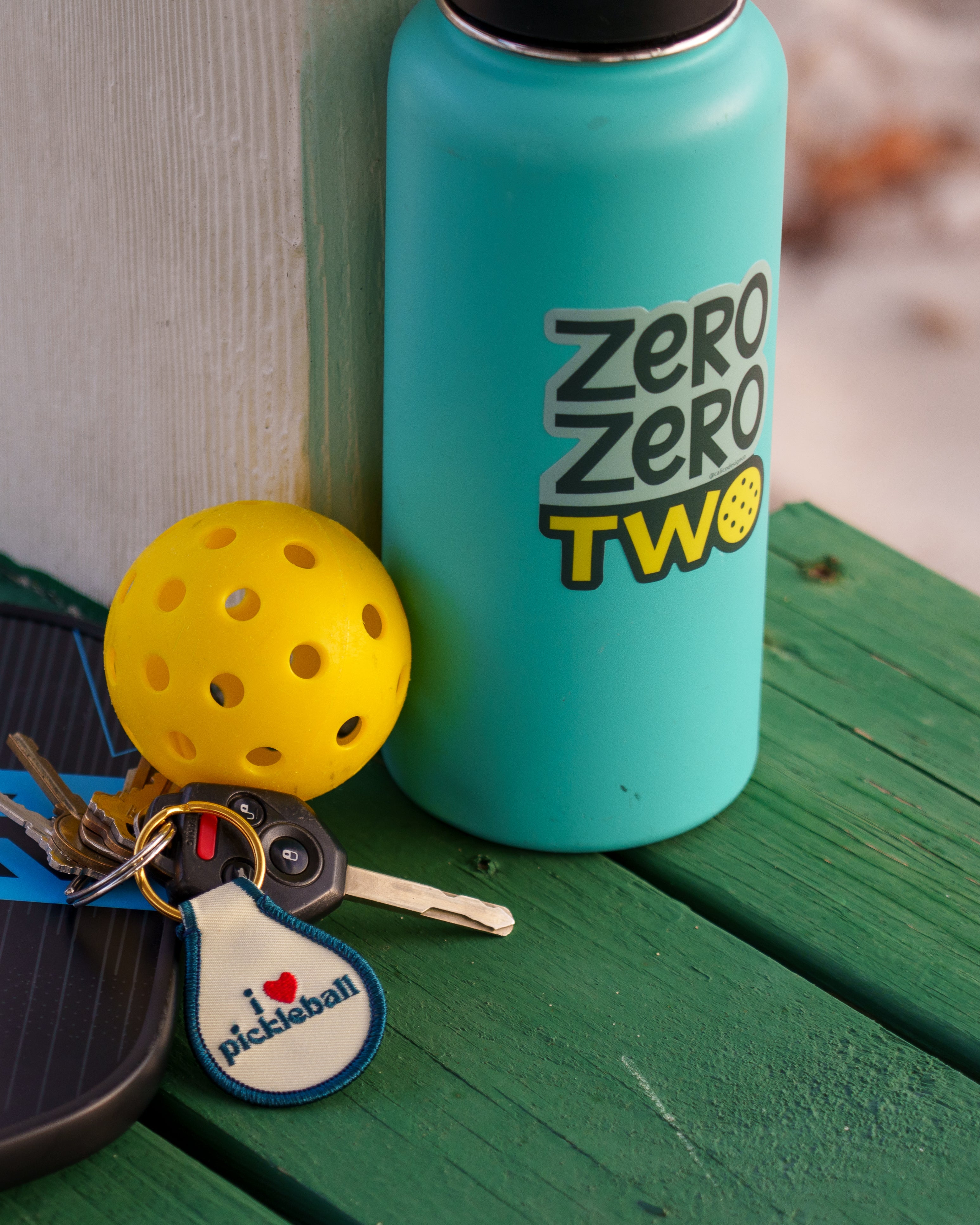 Blue water bottle with a pickleball sticker that says "Zero Zero Two" next to a pickleball, paddle, and keys.