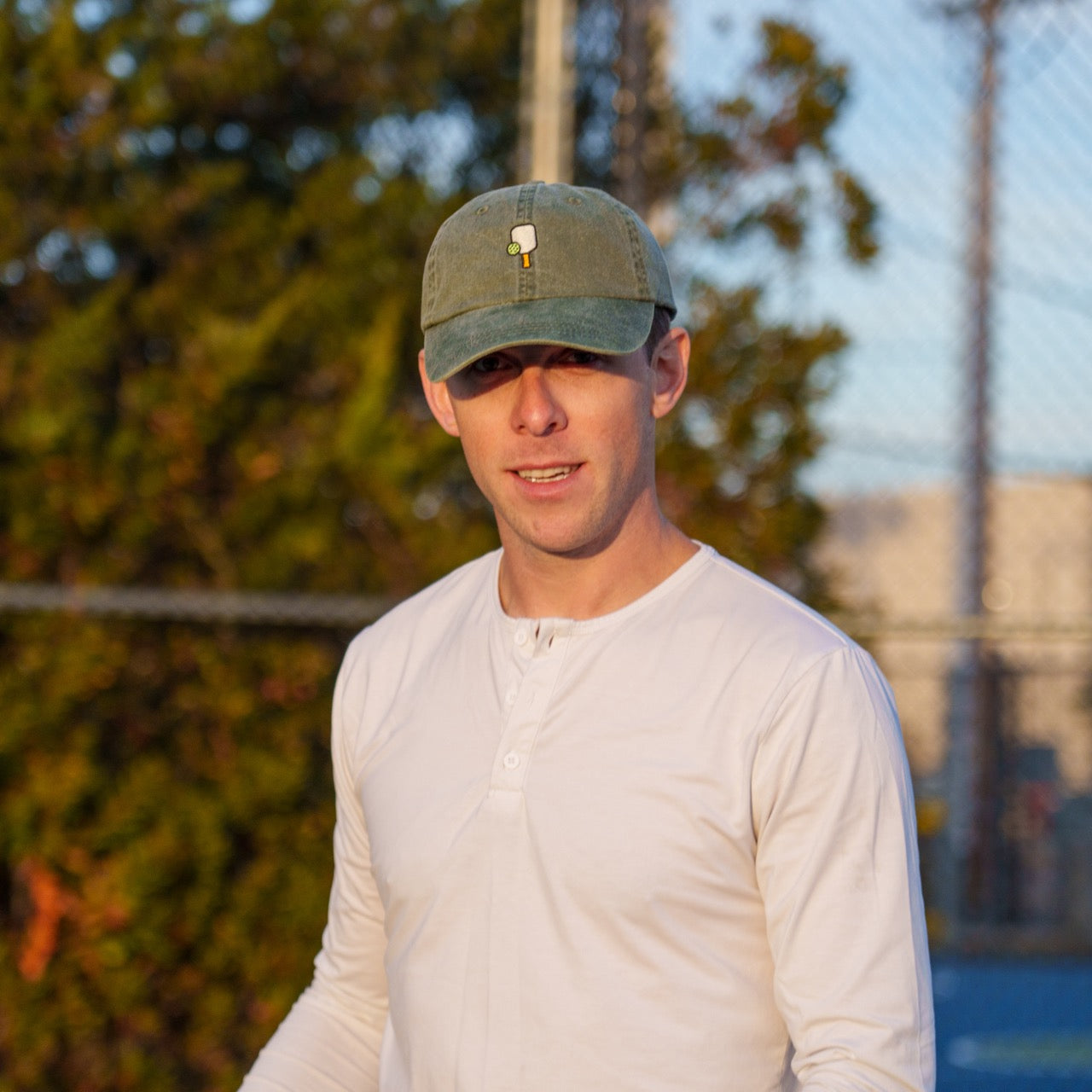 Man on a pickleball court wearing an embroidered baseball hat with a pickleball paddle and ball on the front