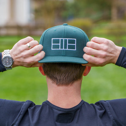 Man wearing a dark green flat brimmed hat with pickleball court lines embroidered on it