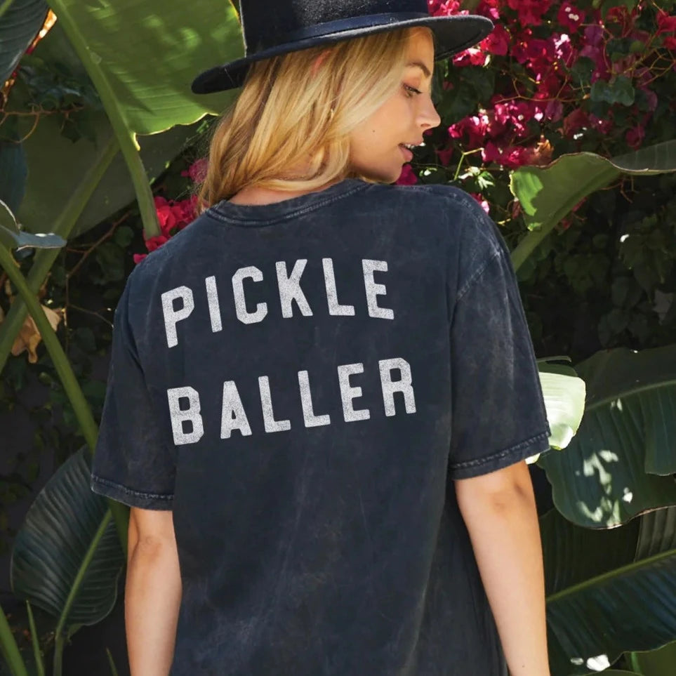 A woman wearing a washed black t-shirt that says &quot;PICKLEBALLER&quot; on the back and a wide brimmed black hat