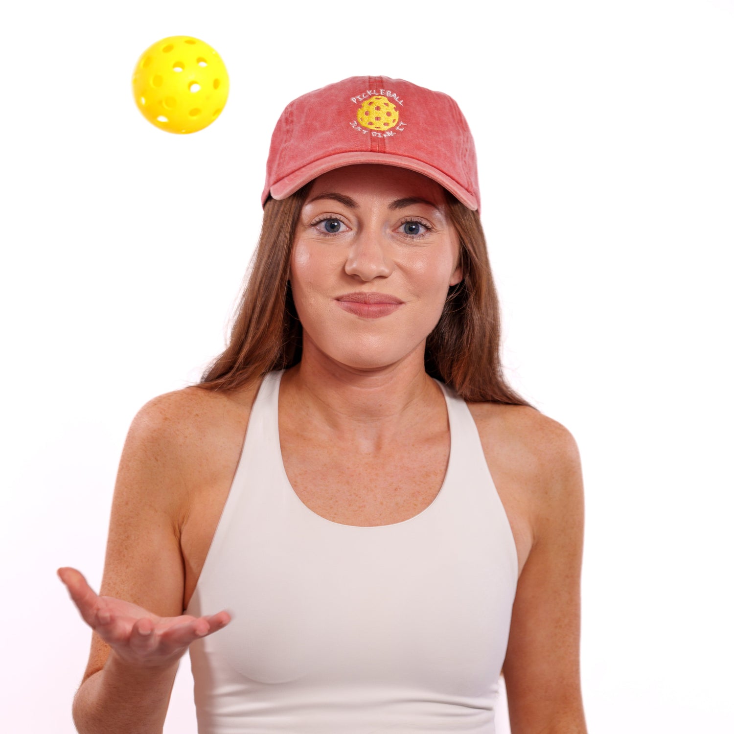 Woman wearing a red hat that says &quot;Pickleball Just Dink It&quot;  throwing up a pickleball in the air