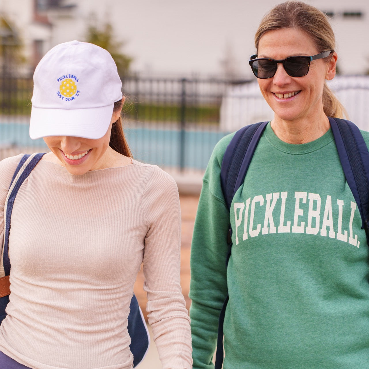 Two women on their way to the pickleball courts, one woman wearing a pickleball sweatshirt and one wearing a white pickleball hat